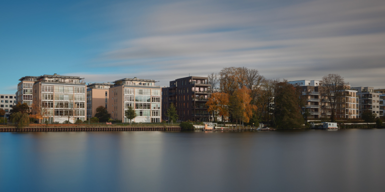 Scenic riverside view of modern buildings reflecting on the calm water under a cloudy sky.