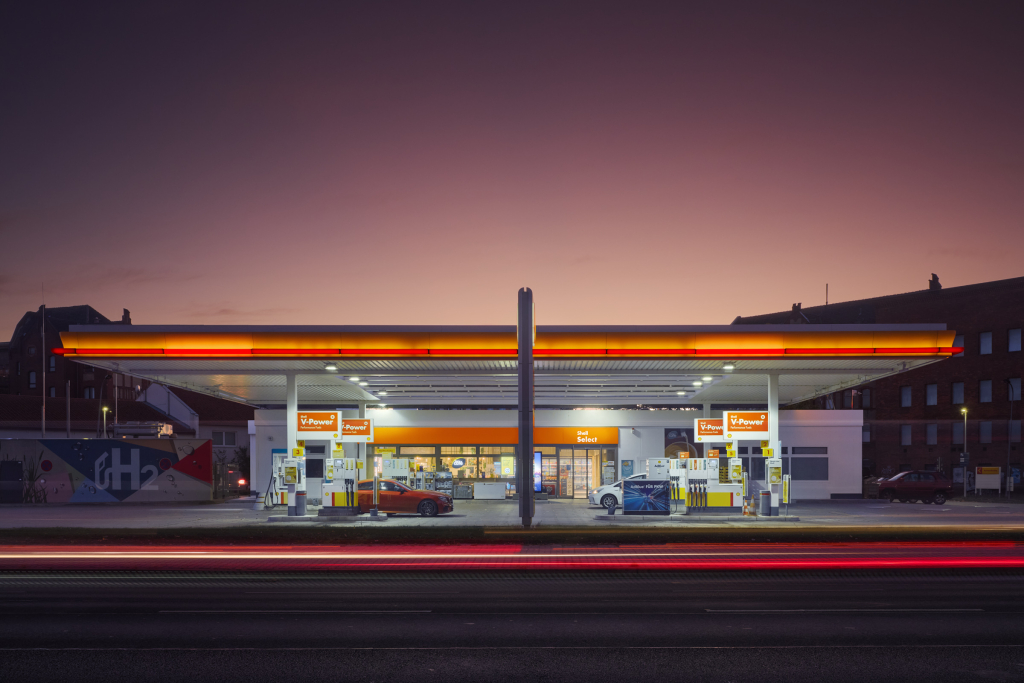 A modern gas station at dusk with illuminated pumps and a sleek canopy, highlighted by vibrant lighting and a colorful sky, creating a cinematic, urban scene.