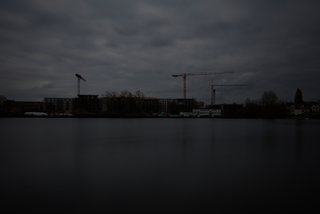 Long exposure of a construction site by a river on an overcast day, significantly underexposed with dark outlines against a gray sky.