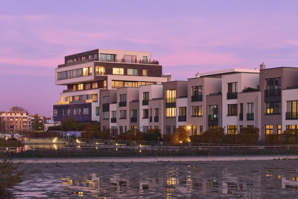 Modern, apartment buildings with warm lights and sleek architecture at dusk, set against a purple-pink sky. Located on the Dahme river near Grünau, Berlin.