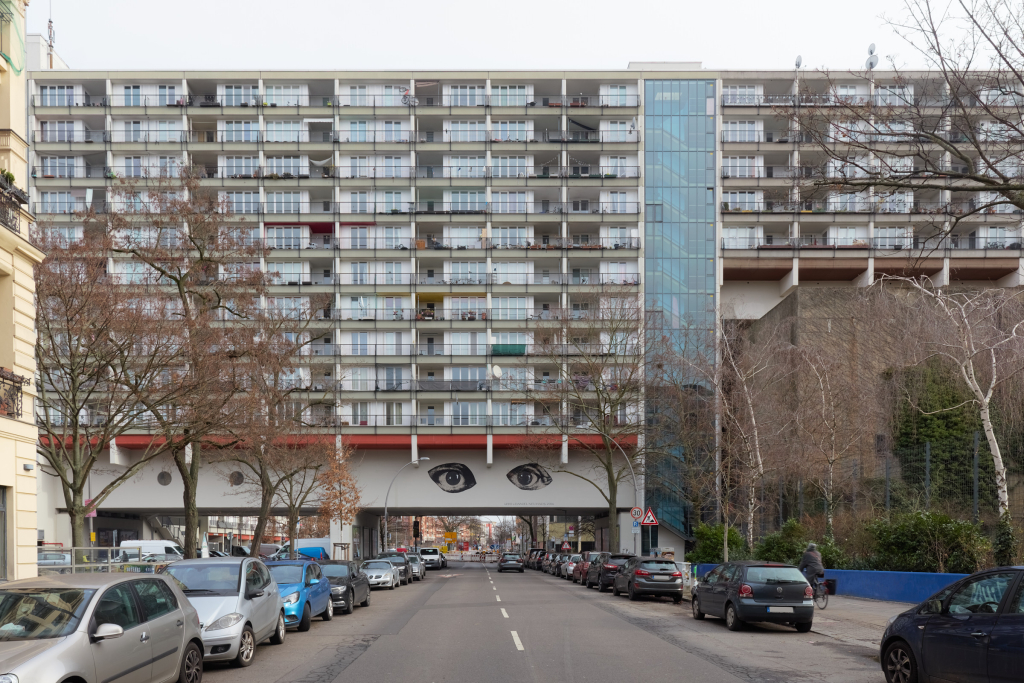 Vorderansicht des Pallasseums in Berlin, mit Blick entlang der Straße auf den massiven Betonbau, auf dem ein Gemälde mit zwei Augen auf dem Bogen zu sehen ist.