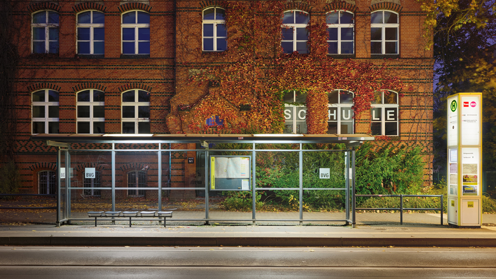 Night view of BVG tram stop near a school in Grünau, Berlin, with autumn leaves and contrasting cool and warm tones.