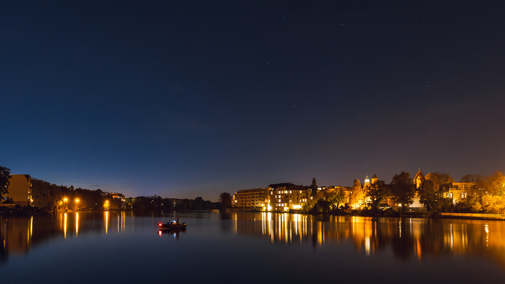 Night long exposure of a man fishing in a boat on the Dahme River with the illuminated cityscape around him.